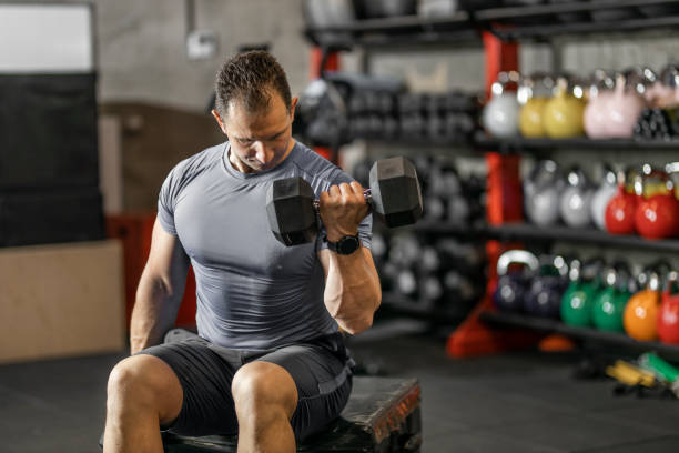 istock Muscular guy in sportswear lifting dumbbell while sitting on bench at cross training gym. Mature athlete using dumbbell during a workout. Strong man under physical exertion pumping up bicep muscle with heavy weight. 1244763920