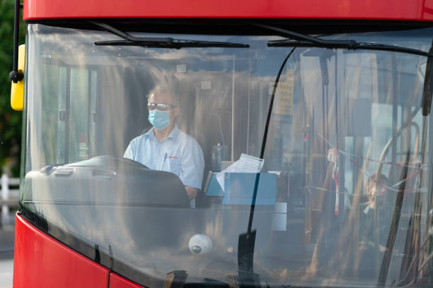 Driver of a double decker red London Bus driving with a face mask on and with security tape for safety during Coronavirus COVID-19 pandemic lockdown on Waterloo Bridge, London, England - 2 A red double decker bus is one of the iconic symbols in London life.  Such buses are now moving to electrical power to mitigate pollution emissions.  This images shows a close up of the driver wearing a face mask during the COVID-19 pandemic. waterloo bridge stock pictures, royalty-free photos & images