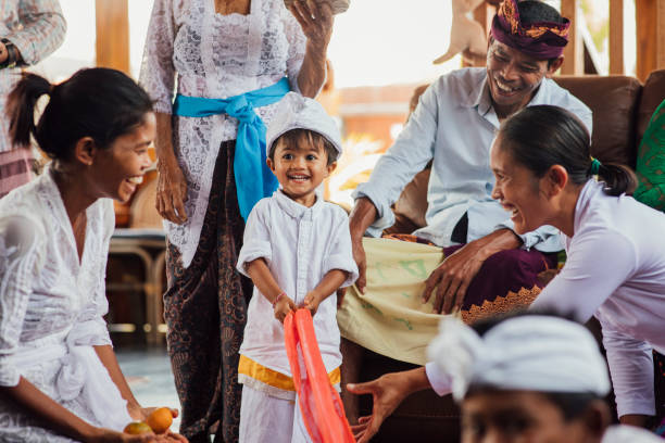 An Indonesian family and their nanny together in the living room at home in Bali, Indonesia. The main focus is the youngest boy holding material as part of a game, while smiling and looking away from the camera. His family are watching him and smiling.
