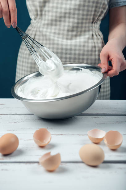 Woman in checkered apron whips egg whites on a white vintage wooden table. Woman in checkered apron whips egg whites on a white vintage wooden table. The process of making meringues. egg white stock pictures, royalty-free photos & images