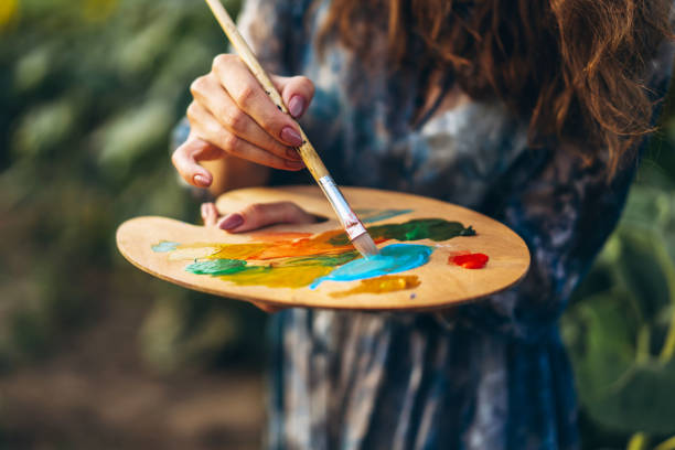 cierre las manos de la artista femenina sosteniendo el pincel y la paleta con pinturas al óleo. fondo borroso con caballete en el campo de girasol - paintbrush palette art paint fotografías e imágenes de stock