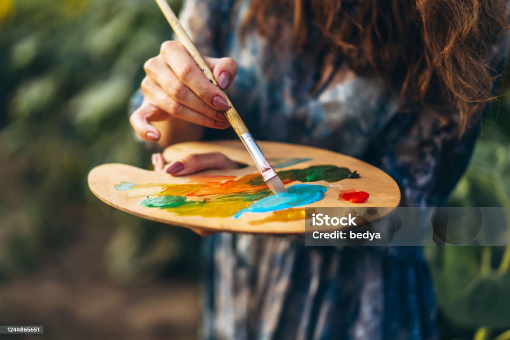 Close up hands of female artist holding brush and palette with oil paints. Blurred background with easel in sunflower field Close up hands of female artist holding brush and palette with oil paints. Blurred background with easel in sunflower field. Painting - Art Product Stock Photo