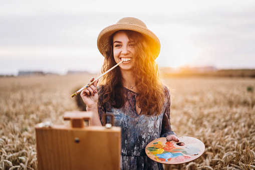 Beautiful girl artist paints in a wheat field. Woman smiles, holds a brush and palette in hands, in front of her stands an easel. Behind her is orange sunlight.