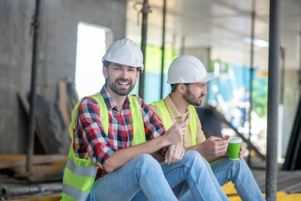 istock Building workers in yellow vests and helmets sitting, having coffee with sandwiches, laughing 1244893336