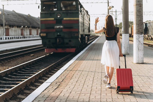 Female brunette traveler with red suitcase in white skirt waiting for a train and waving hand to the train on raiway station