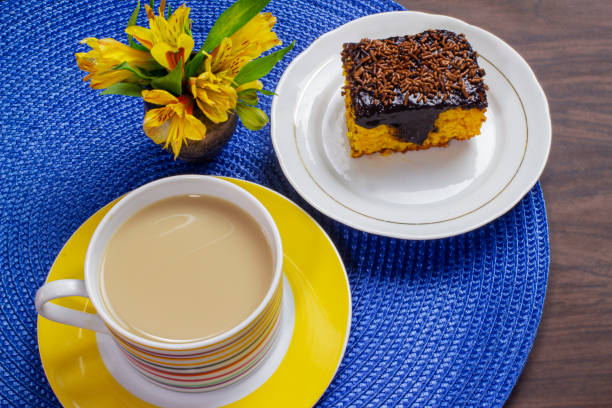 bolo de cenoura com cobertura de chocolate e xícara de leite com café em mesa de madeira decorada com uma flor amarela. - cake yellow sweet food banquet - fotografias e filmes do acervo