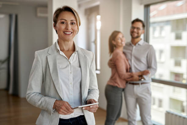 Portrait of happy real estate agent with a couple in the background. Portrait of female real estate agent looking at the camera while happy couple in standing in the background. estate agent stock pictures, royalty-free photos & images