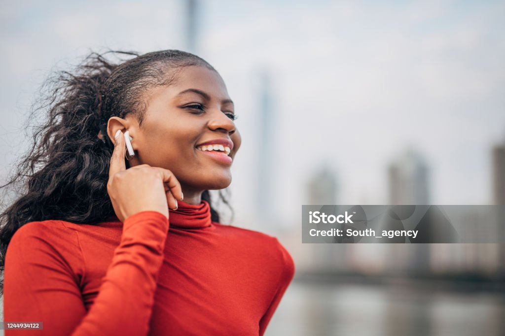 One woman listening music on wireless headphones by the river One beautiful young woman, black woman, standing downtown by the river and listening music on wireless headphones. Wireless In-ear Headphones Stock Photo