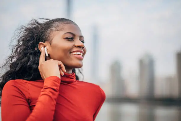 One beautiful young woman, black woman, standing downtown by the river and listening music on wireless headphones.