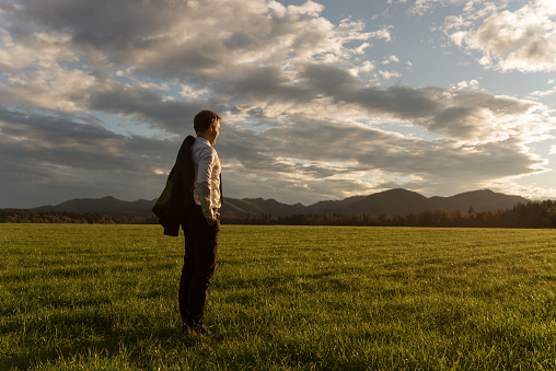 Young businessman with his suit jacket over the shoulder, standing in beautiful green meadow under dramatic evening sky gazing into the distance.