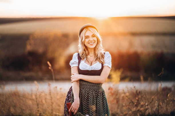 Pretty blonde girl in dirndl, standing outdoors in the field, holding bouquet of a field flowers. stock photo