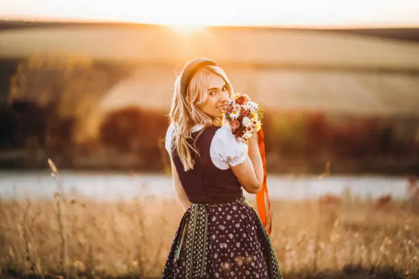 Pretty blonde girl in dirndl, traditional festival dress, standing outdoors in the field at sunset time, holding bouquet of a field flowers. Beer Fest, St. PatrickÃ¢Â€Â™s day, international beer day concept