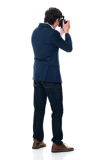 Rear view of aged 20-29 years old with short hair caucasian male photographer standing in front of white background wearing blazer who is photographing and holding camera