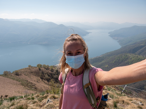 Selfie of woman hiking outdoor wearing a medical face mask to prevent COVID-19 and social distancing from people. Female with facial mask hiking enjoying nature, coronavirus pandemic protection