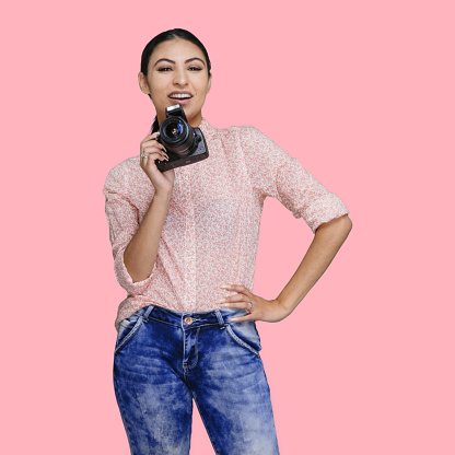 Waist up of aged 20-29 years old who is beautiful with black hair latin american and hispanic ethnicity female photographer standing in front of colored background wearing jeans who is happy who is photographing and holding camera