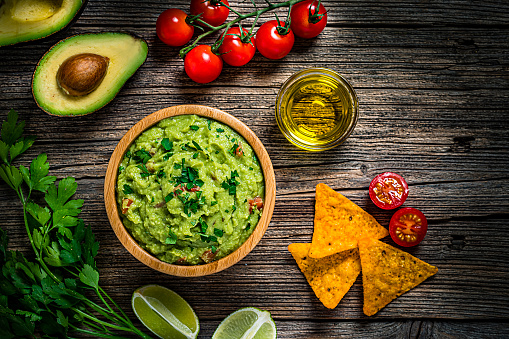 Mexican food: guacamole dipping sauce in a bowl with tortilla chips shot from above on rustic wooden table. Ingredients for preparing guacamole are all around the bowl and includes ripe avocados, tomatoes, lime, cilantro and salt. Predominant color is green. High resolution 42Mp studio digital capture taken with Sony A7rII and Sony FE 90mm f2.8 macro G OSS lens