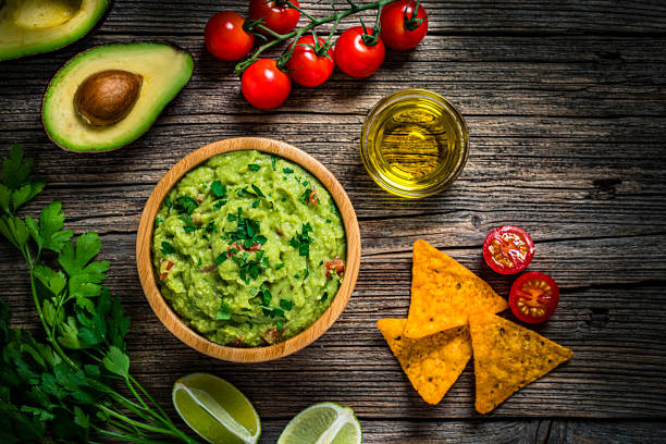 Guacamole and chips on rustic wooden table Mexican food: guacamole dipping sauce in a bowl with tortilla chips shot from above on rustic wooden table. Ingredients for preparing guacamole are all around the bowl and includes ripe avocados, tomatoes, lime, cilantro and salt. Predominant color is green. High resolution 42Mp studio digital capture taken with Sony A7rII and Sony FE 90mm f2.8 macro G OSS lens guacamole stock pictures, royalty-free photos & images