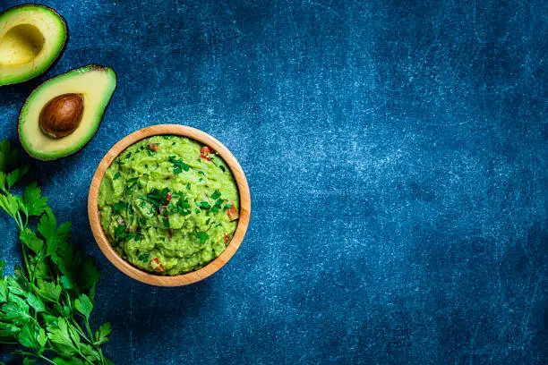 Mexican food: guacamole dipping sauce in a bowl shot from above on bluish tint table. Some ingredients like ripe avocados, lime and cilantro are beside the bowl. The composition is at the left of an horizontal frame leaving useful copy space for text and/or logo at the right. Predominant colors are green and blue. High resolution 42Mp studio digital capture taken with Sony A7rII and Sony FE 90mm f2.8 macro G OSS lens