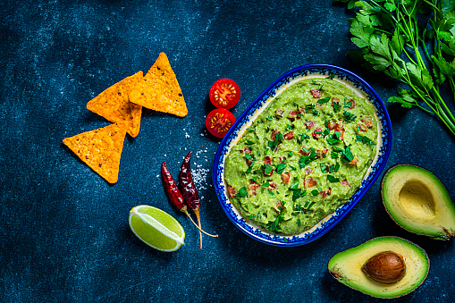 Bowl of delicious guacamole, nachos chips and ingredients on grey table, flat lay. Space for text