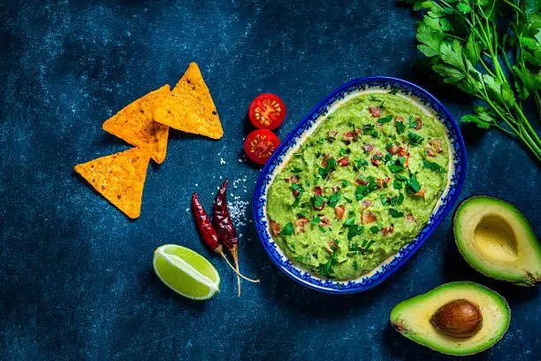 Mexican food: guacamole dipping sauce in a bowl with tortilla chips shot from above on abstract bluish tint table. Ingredients for preparing guacamole are all around the bowl and includes ripe avocados, tomatoes, lime, cilantro and salt. Predominant colors are green and blue. High resolution 42Mp studio digital capture taken with Sony A7rII and Sony FE 90mm f2.8 macro G OSS lens