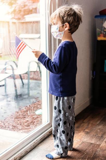 bambino che guarda attraverso una finestra usando una maschera protettiva con in mano una bandiera degli stati uniti - depression sadness usa american flag foto e immagini stock