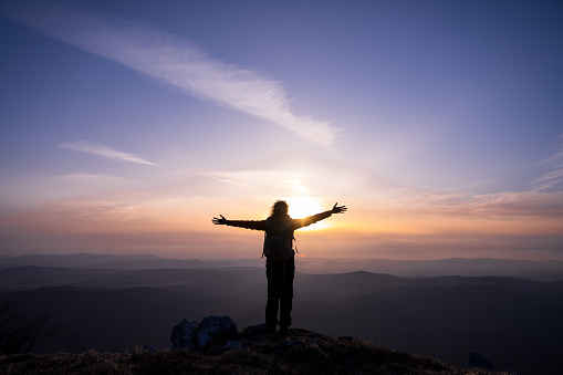 Rear view of silhouetted woman with arms outstretched standing on mountain during sunset.