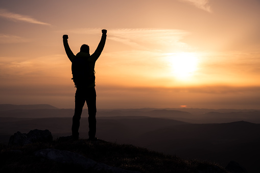 Rear view of silhouetted man with arms raised standing on mountain against sky during sunset.