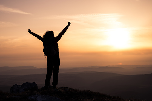 Rear view of silhouetted woman with arms raised standing on mountain during sunset.