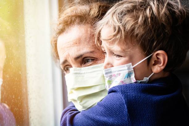 istock Mature woman posing with her son, both with protective masks, very sad looking through window worried about Covid-19 lockdown 1245181743