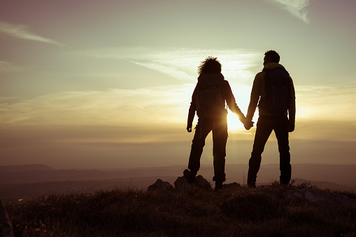 Rear view of silhouetted man and woman holding hands while standing on mountain during sunset.