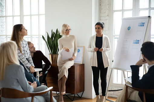 Young focused indian female speaker coach standing near whiteboard, giving marketing research results presentation or training to focused diverse colleagues employees businesspeople in modern office.