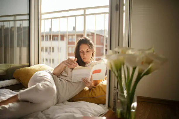 Young woman lying on bed next to the open balcony window and reading a book. Apartment has a nice view and beautiful light at morning. She wears pajamas and looks serene and beautiful.