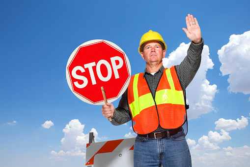 A low angle view of a highway worker holding up a stop sign while using his other hand to signal to drivers to please stop as he stands in front of a cloud filled sky.