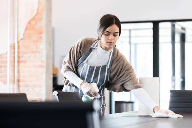 Young woman uses protective gloves and disinfecting spray to clean The young woman uses protective gloves, a disinfecting spray, and paper towels to clean a conference table at work. paper towel stock pictures, royalty-free photos & images