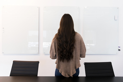 Rear view of a female creative professional standing in front of three blank whiteboards. She is considering ideas for a project.