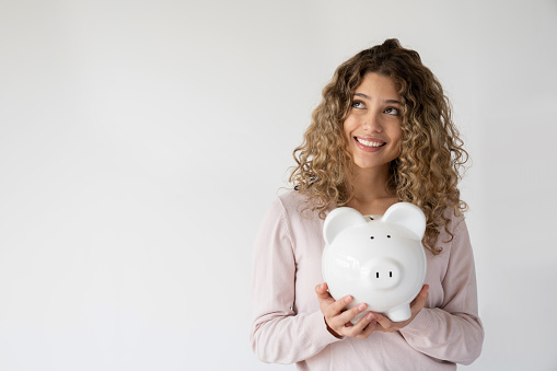 Portrait of a happy woman holding a piggybank and thinking about how to spend her savings while smiling over a white background