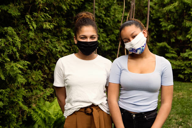 Teenage sisters smiling behing stylish protective mask. Beautiful teenage sisters smiling behind their stylish protective black mask. They are mixed-race and looking at the camera. Nature in background. Horizontal outdoors waist up shot with copy space. teenage girls pretty smile looking at camera waist up stock pictures, royalty-free photos & images