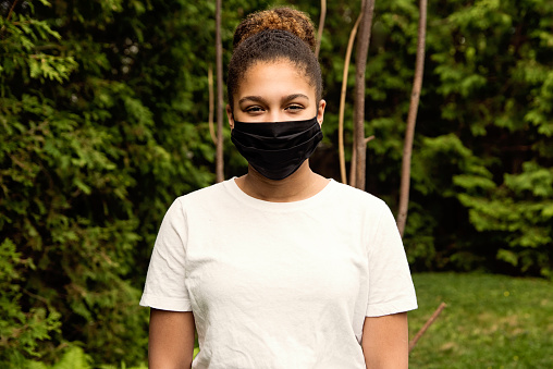 Beautiful teenage girl smiling behind her stylish protective black mask. She his mixed-race and is looking at the camera. Nature in background. Horizontal outdoors waist up shot with copy space.