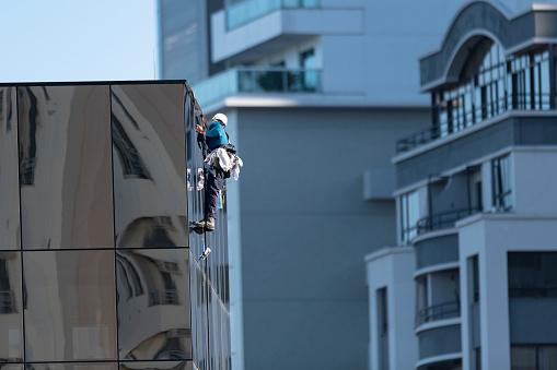 Image of a young skyscraper window washer wearing safety equipments and cleaning a glass curtain wall building facade