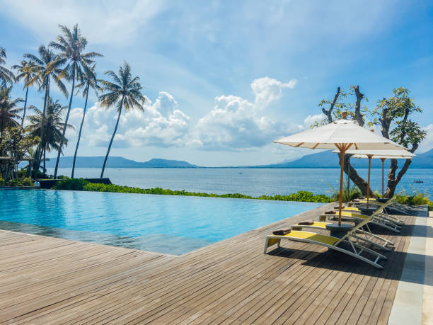 Beautiful tropical swimming pool in hotel or resort with umbrella, coconuts tree sun-loungers, palm trees with infinity pool view, ocean and mountain background. Beautiful tropical swimming pool in hotel or resort with umbrella, coconuts tree sun-loungers, palm trees with infinity pool view, ocean and mountain background. jawa timur stock pictures, royalty-free photos & images