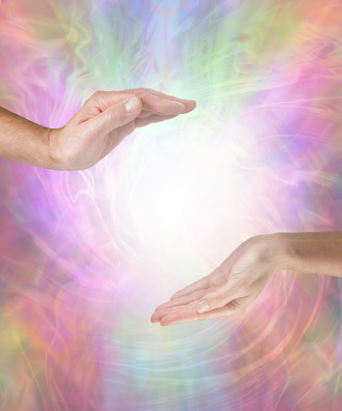 male hand opposite female hand with a white light orb in between against a multicoloured flowing energy field background with copy space