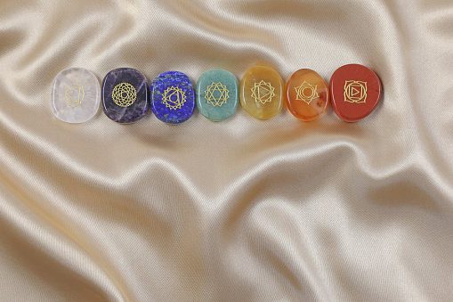 magenta, indigo, blue, green, yellow, orange and red polished flat gem stones etched with 7 chakra symbols on a gold silk material background with copy space