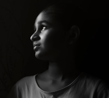 Indoor black and white, low key, close-up portrait of an Asian/Indian sad, innocent girl looking out through the window and thinking soothing with a blank expression on her face.