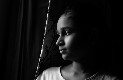 Indoor black and white, low key, close-up portrait of an Asian/Indian sad, serene girl looking out of the window under natural light and thinking soothing with a blank expression.