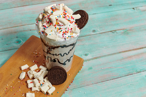 still life with a beautiful milkshake in a transparent glass. milk drink is very sweet and delicious with marshmallows, cookies, whipped cream and colorful sprinkles on a light blue wooden background