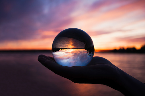 Beautiful scenic view through lens of crystal ball on water horizon at summer sunset. Clouds reflection in sphere. Palm of hand holding globe in outdoor.