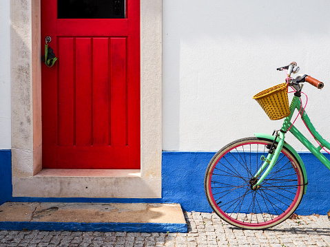 An old model bicycle with a lady's wicker basket parked in front of a house with red doors and windows