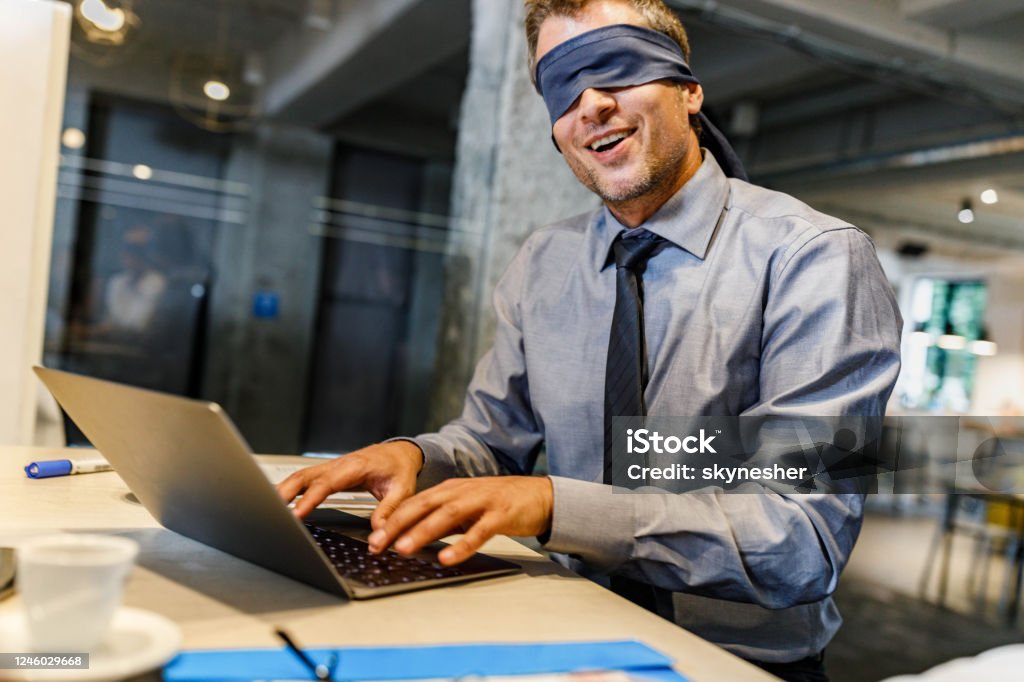 Blindfolded businessman unable to see the reality over computer in the office. Happy blindfolded businessman cannot see the real picture while using computer in the office. Blindfold Stock Photo