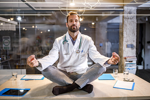 Male doctor doing Yoga breathing exercises in Lotus position on the table in the office. The view is through glass.