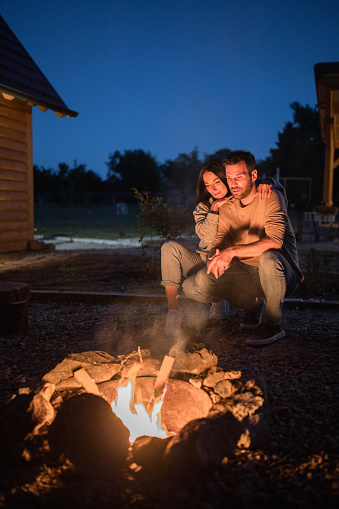 Loving couple crouching by the campfire during the night in their backyard. Copy space.